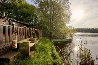 a cabin by the water with a bench and a lake at Buzzard Lodge with Hot-tub in Carlisle