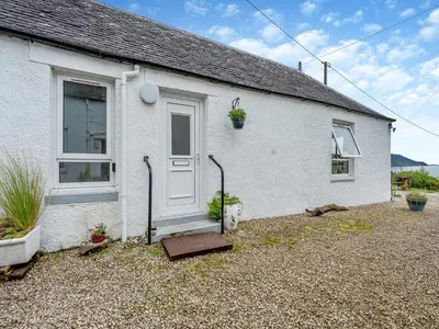 a white house with a door and a cat sitting outside at Carraig Dhubh Cottage - Uk47123 in Whiting Bay