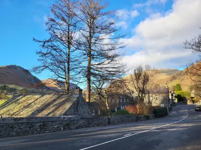 a street with a stone wall and trees on a hill at Holly Cottage Grasmere in Grasmere