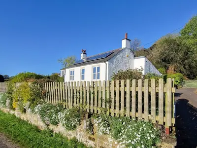 a white house behind a wooden fence with flowers at Bankswood Cottage in Ruthwell