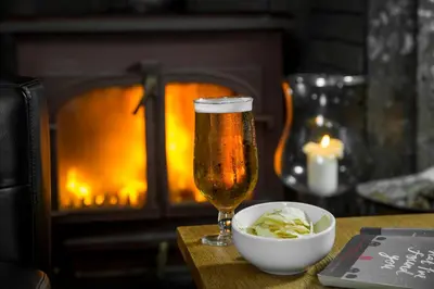 a glass of beer and a bowl of chips on a table with a fireplace at The Greenhill Hotel in Wigton