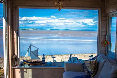 a living room with a large window looking out at a beach at Sunset Retreat Lodge with Hot Tub in Carsethorn