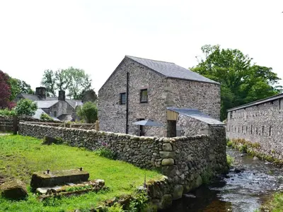 a stone house with a stone wall next to a river at Shepherds Cottage in Corney