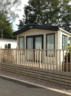 a small house with a wooden fence on the side of the road at Woodpecker lodge, Camelot Holiday Park, CA6 5SZ in Carlisle