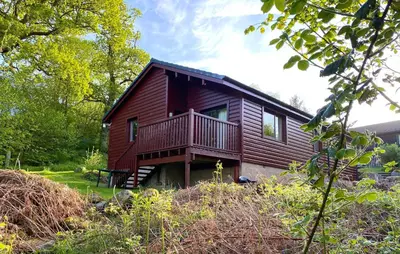 a red cabin with a deck in the woods at Beautiful 3BR Lodge - Stunning Beach and Golf Location - Pass the Keys in Dalbeattie